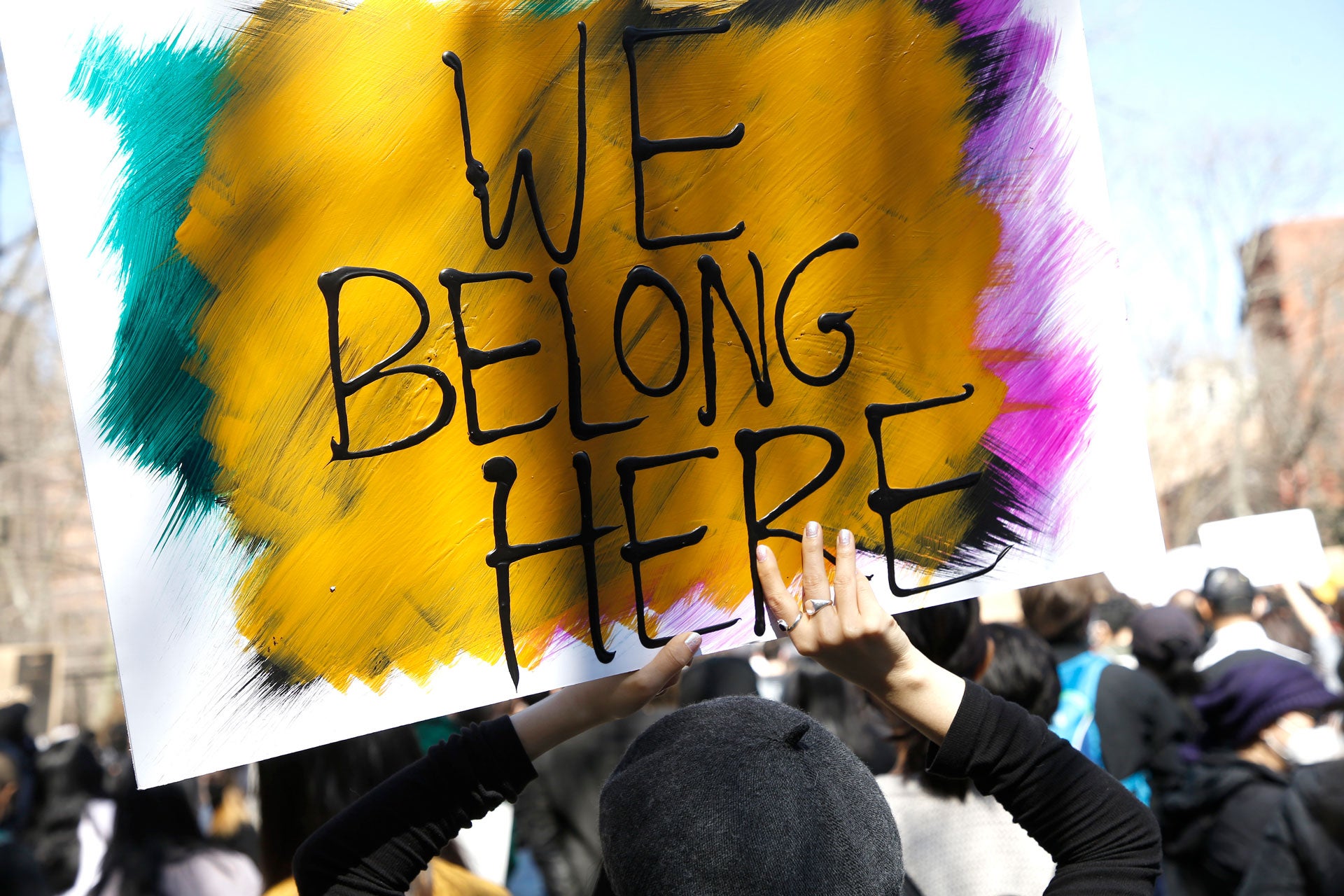 People demonstrate at a rally in Chinatown, New York City. Someone is pictured holding a painted sign reading "We Belong Here" with yellow, blue, pink, and black colors..