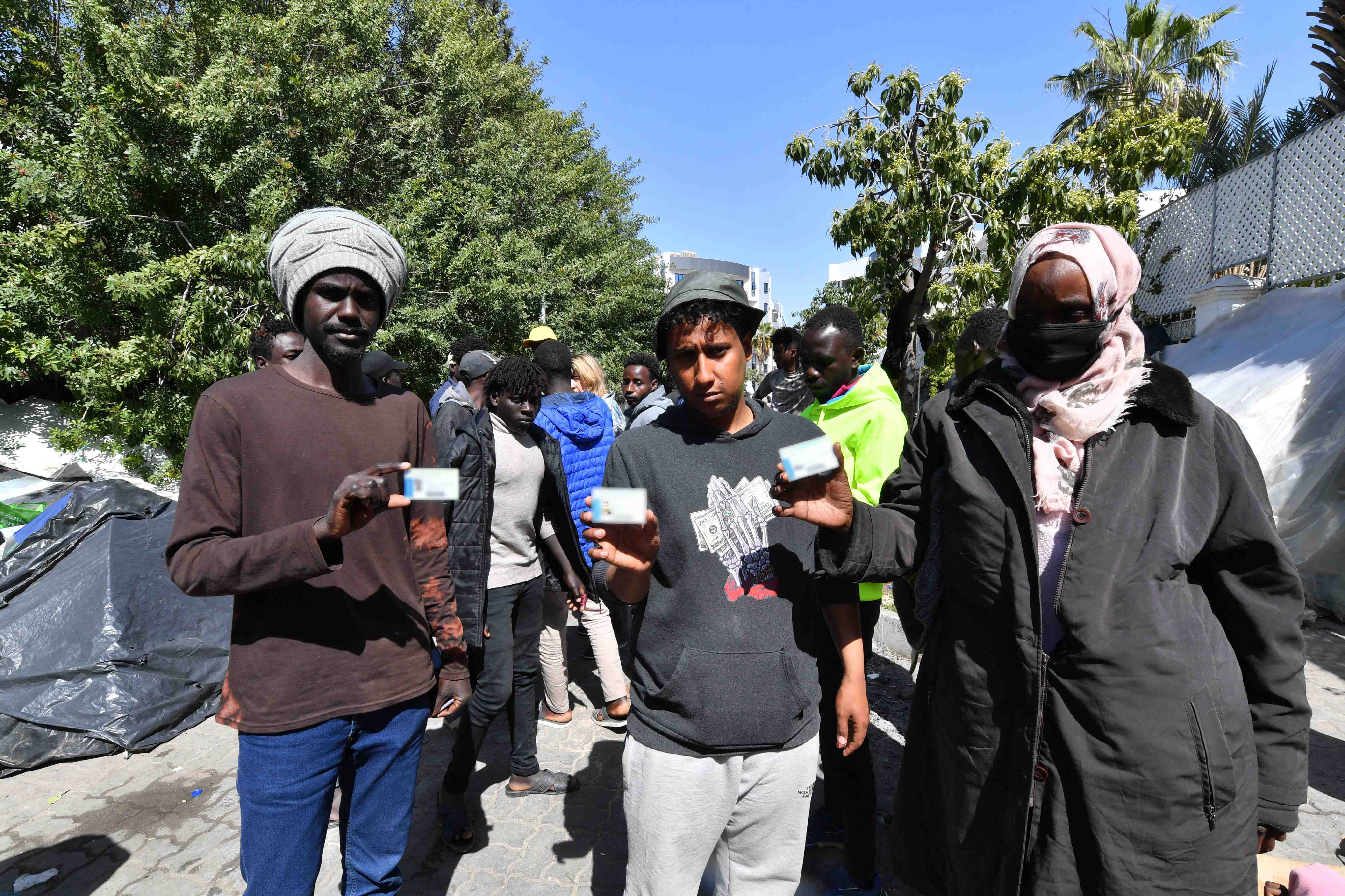 Sudanese and Yemeni men show their refugee or asylum seeker cards on April 12, 2023 outside the International Organization for Migration (IOM) office in Tunis, after Tunisian police on April 11 used force and tear gas to disperse refugees, asylum seekers, and migrants and dismantled their makeshift camp in front of the nearby UN Refugee Agency (UNHCR) office in Tunis. © 2023 Fethi Belaid/AFP via Getty Images