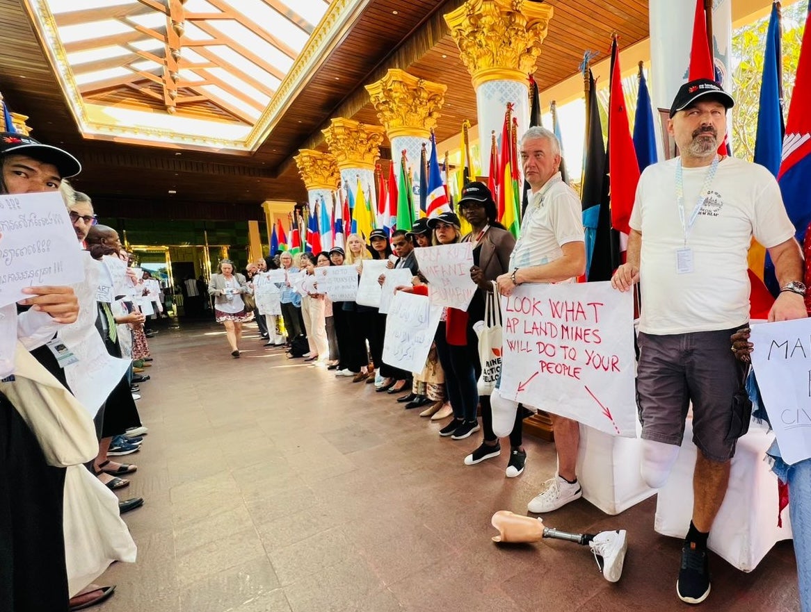 People protest in a corridor with two men with amputated legs hold a sign that says "Look what AP landmines will do to your people."