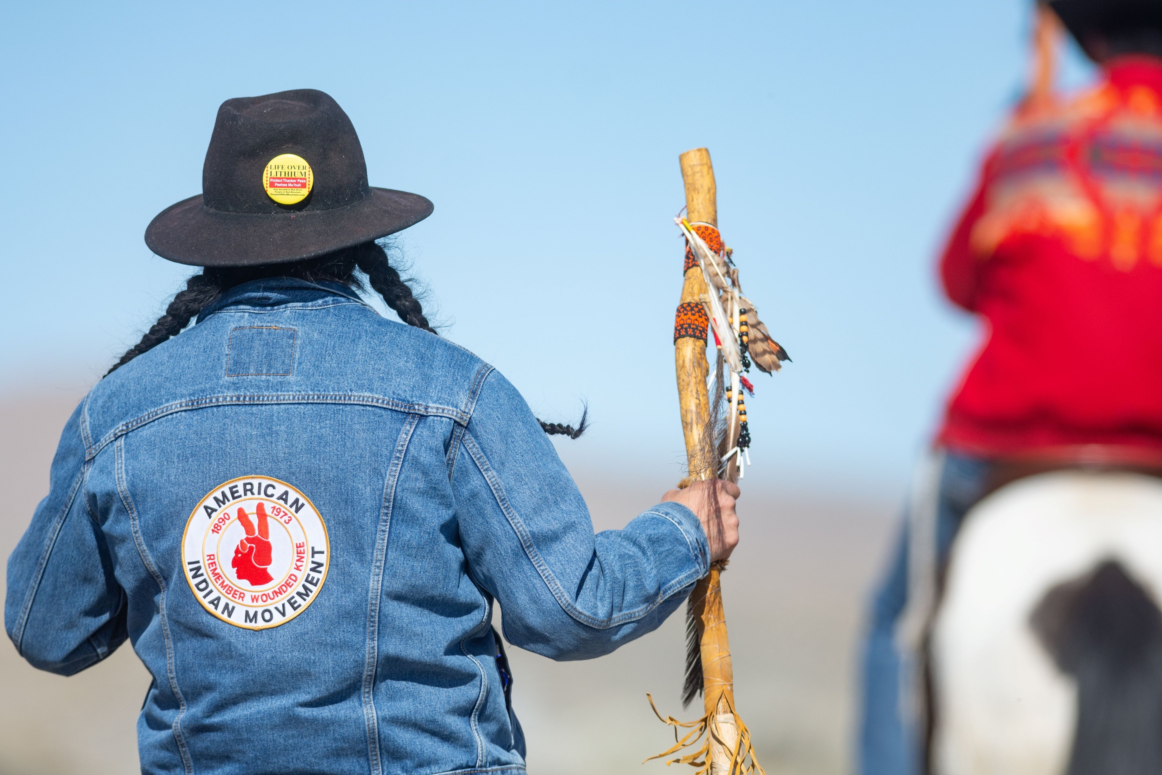 A Numu/Nuwu and Newe individual with a ceremonial staff and a pin reading “Life over Lithium”