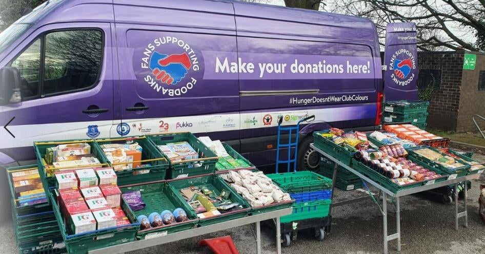 A mobile food pantry with food collected by Fans Supporting Foodbanks and other local anti-poverty groups set up at at St. Mary’s Millennium Centre, Liverpool, UK. Photo shows a purple van with cases of food set up in front of it.