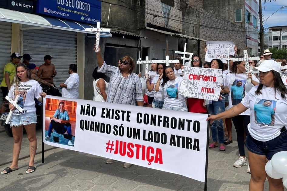  The image shows a group of people participating in a protest. The protesters hold banners and wear t-shirts with a person's face. One prominent banner read, in Portuguese, "There is no confrontation when only one side shoots" with the word "JUSTICE" emphasized below. 