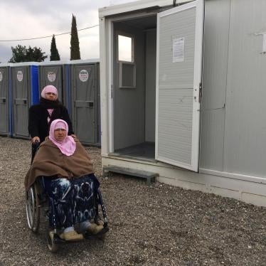 Naima, 70, an older woman with a disability from Aleppo, Syria, with her daughter Hasne, in front of the shower area in Cherso camp, Thessaloniki. The showers are not accessible for people who use a wheelchair.