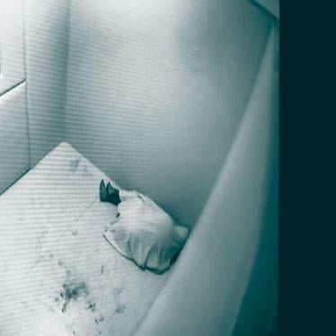 A person lying on the floor of a prison cell, seen through a CCTV camera.